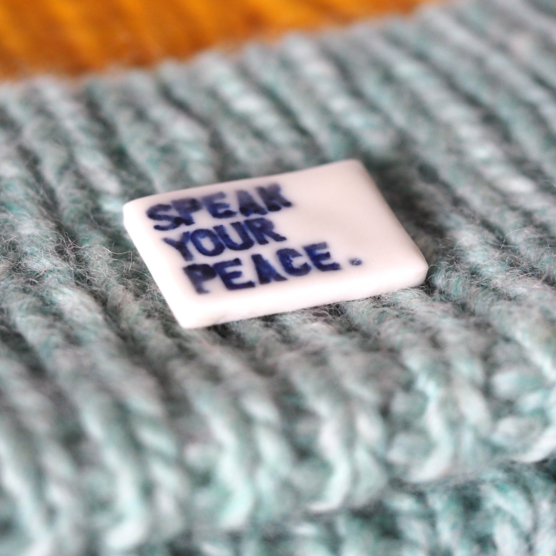 Small rectangular tile with 'SPEAK YOUR PEACE.' text on a textured fabric background